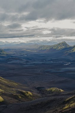 Dolambaçlı bir nehir, karanlık bir volkanik arazi ve yeşil yosun vadisinde akar. Yuvarlanan tepeler, uzak dağlar ve bulutlu bir gökyüzü tarafından çerçevelenir..