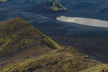 İzlanda 'nın engebeli volkanik arazisinde yosun kaplı tepeler, yansıtıcı bir göl ve dar bir yamaçta yürüyüşçüler bölgenin uzak ölçeğini vurguluyor..
