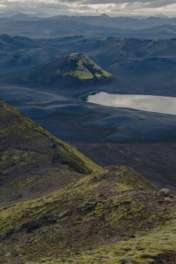 İzlanda 'daki engebeli volkanik arazi, yosun kaplı bir koni, yansıtıcı bir göl, uzak dağlar ve ön taraftaki yamaçlardan geçen yürüyüşçüler..