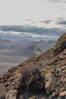 Önünde pürüzlü kayalar olan engebeli volkanik arazi, İzlanda 'da bulutlu bir gökyüzünün altında yosun kaplı tepe, çorak arazi ve uzak tepeler..