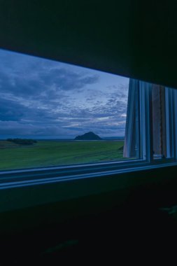 Alacakaranlıkta sakin çimenli bir manzara açık bir pencere tarafından çerçevelenir, bulutlu bir gökyüzünün altında açık bir tepe yumuşak mavi tonlarla.