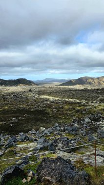 İzlanda 'da bulutlu bir gökyüzünün altında kayan tepeler ve uzak dağlarla, engebeli volkanik kayalar ve yosunlar sahneyi kapladı..
