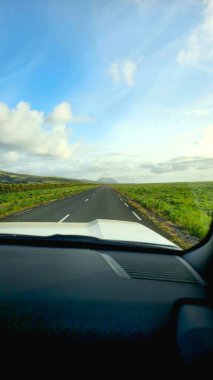 Asfaltlı bir yol, parçalı bulutlu bir gökyüzünün altında yeşil tarlalarla çevrili olan mesafeye uzanır. Arka planda bir dağ yükseliyor, bir araçtan görülüyor..