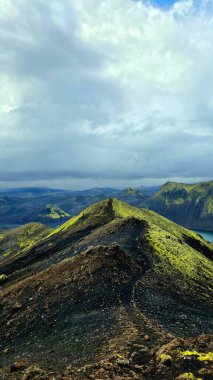 İzlanda 'daki engebeli volkanik sırt yeşil yosun, kayalık arazi ve soluk bir iz. Sarp dağlar ve yuvarlanan tepeler bulutlu bir gökyüzünün altında belirir..