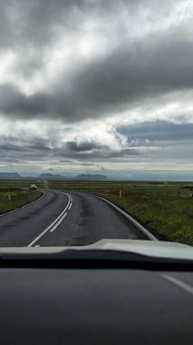 Beyaz şeritli dolambaçlı bir yol bulutlu bir gökyüzünün altındaki yeşil çayırları kesiyor. Ufukta dağlar görünüyor, içinde bir araba görünüyor..
