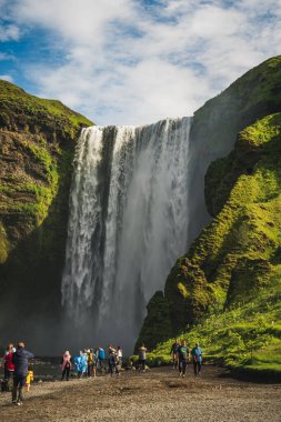 Skogafoss şelaleleri, parçalı bulutlu bir gökyüzünün altındaki yeşil uçurumların 60 metre aralığında. Üsse gelen ziyaretçiler ölçeği ve sisli çevreyi vurguluyorlar.