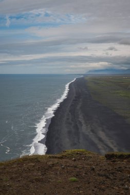 Reynisfjara 'nın İzlanda' daki siyah kumsal sahili, beyaz okyanus dalgaları, kayalık arazi, yeşil tarlalar ve bulutlu gökyüzünün altındaki uzak dağlar..
