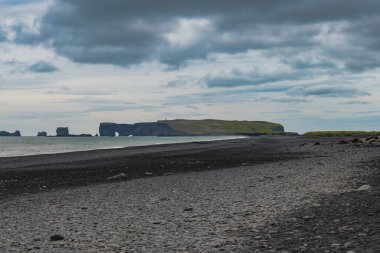 Pürüzsüz çakıl taşlarıyla siyah bir kumsal ve uzakta Dyrholaey hortumu, bulutlu gökyüzünün altında ikonik kemeri ve engebeli uçurumları barındırıyor..