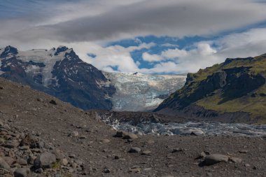 Mavimsi beyaz renklere sahip bir buzul volkanik tepeler ve İzlanda 'nın yosunlu yosunlu yamaçları arasında uzanır. Kayalık bir ön planı ve parçalı bulutlu gökyüzü vardır..