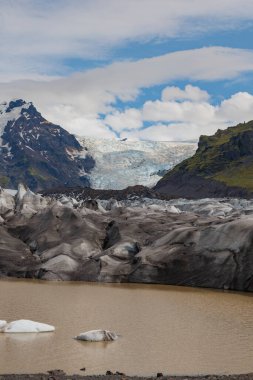 İzlanda 'daki bir buzul engebeli zirvelerden yeşil yamaçlar ve yukarıda bulutlu bir gökyüzü tarafından çerçevelenmiş yüzen buz kütleleriyle çamurlu bir lagüne uzanır..