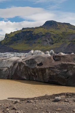 Volkanik küller, çamurlu bir buzul gölü ve İzlanda 'nın engebeli arazisinde parçalı bulutlu bir gökyüzünün altında yeşil kaplı bir dağ..