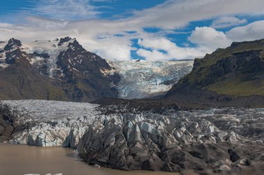 İzlanda 'daki bir buzul, karlı zirveleri olan karanlık dağlarla çevrili çamurlu bir buzul gölüne doğru uzanır. Arka planda bulutlu bir gökyüzünün altında büyük bir buz tabakası yatıyor..
