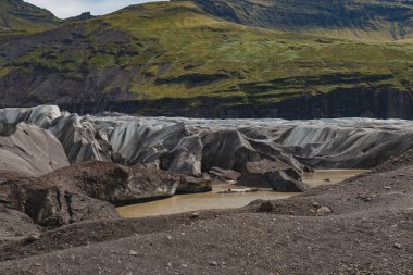 Gri çizgili buzullar, kayalık bir ön planla, çamurlu bir dereyle ve İzlanda 'nın engebeli arazisindeki dik yosun kaplı tepelerle çelişiyor..