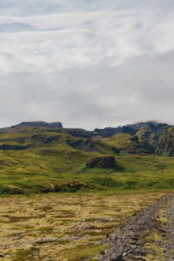 Yuvarlanan yeşil tepeler yosun kaplı arazi ve kayalık çıkıntılar, ön planda bir çakıl yolu ve bulutlu bir gökyüzünün altındaki sarp dağ zirveleri..