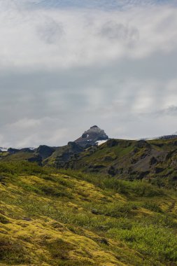 Karla kaplı zirvesi, sivri uçurumları ve parçalı bulutlu bir gökyüzünün altındaki canlı yosunlarıyla İzlanda 'nın engebeli manzarası..