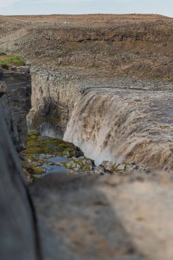 Dettifoss yükselen sis ile engebeli bir uçurumdan atlıyor. Rocky arazisi, seyrek bitki örtüsü ve uzak figürler şelalenin geniş ölçeğini ön plana çıkarıyor..