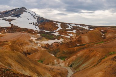 İzlanda 'daki Kerlingarfjoll sıradağlarında turuncu ve kahverengi rhyolite tepeler, kar parçaları, dolambaçlı bir dere ve bulutlu bir gökyüzü bulunur..