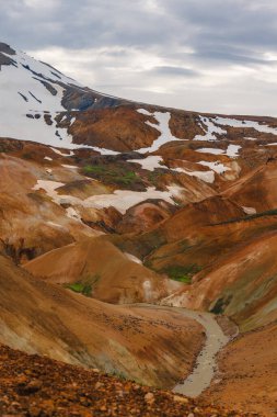 Canlı turuncu, kırmızı ve kahverengi rhyolite dağları, kar parçaları ve Kerlingarfjoll, İzlanda 'da bulutlu bir gökyüzünün altında dolambaçlı bir nehir..