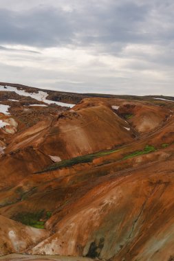 Canlı turuncu, kırmızı ve kahverengi rhyolite tepeleri Kerlingarfjoll, İzlanda 'da dağınık kar yamaları ile seyrek bitki örtüsü ile bulutlu bir gökyüzü altında.