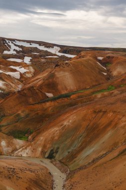 Canlı turuncu, kırmızı ve kahverengi rhyolite tepeleri dağınık kar yamaları, dolambaçlı bir dere ve Kerlingarfjoll, İzlanda 'da bulutlu bir gökyüzü.