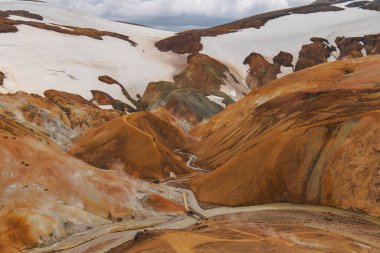 Turuncu, sarı ve kahverengi renkli Rhyolite dağları kar yamaçları, dolambaçlı patikalar ve İzlanda, Landmannalaugar 'da bulutlu bir gökyüzü altında dereler..