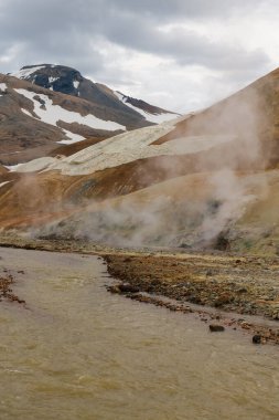 İzlanda 'da çamurlu bir nehrin yanında buharlı bacalar yükseliyor. Etrafı çok renkli rhyolite tepeleri, kar bantları ve arka plandaki engebeli dağlarla çevrili..