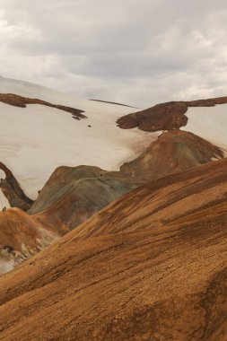 İzlanda 'nın dağlık bölgelerindeki bulutlu bir gökyüzünün altında, beyaz kar parçalarıyla zıt kırmızı, turuncu ve yeşil tonlara sahip çok renkli ryolite tepeleri.