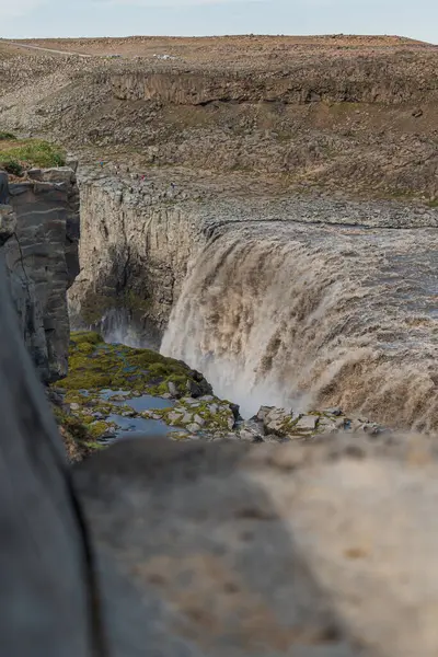 Dettifoss yükselen sis ile engebeli bir uçurumdan atlıyor. Rocky arazisi, seyrek bitki örtüsü ve uzak figürler şelalenin geniş ölçeğini ön plana çıkarıyor..