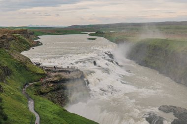 Gulfoss şelalesi yükselen sis ile engebeli bir vadiye akar. Bir izleme platformu, dolambaçlı bir yol, yeşil tepeler ve bulutlu bir gökyüzü sahneyi tamamlıyor..