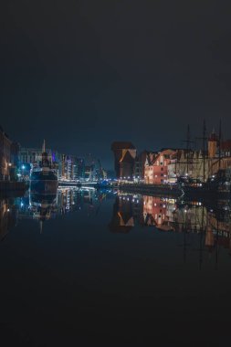 Polonya 'nın Gdansk kentindeki Motlawa Nehri' nin sakin gece sahnesinde aydınlanmış Zuraw vinci, tarihi cepheler, modern yapılar ve sakin yansımalar yer alıyor..