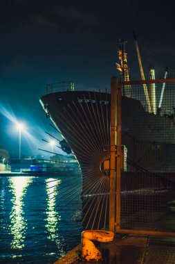 Büyük bir gemi, Polonya 'nın Gdansk kentindeki çitli bir bölgeye gece yanaştı. Sokak lambaları durgun suyu yansıtıyor, endüstriyel ve deniz elementleri görünür..