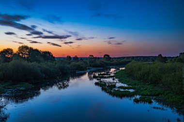 Sakin bir nehir, alacakaranlıkta yoğun bir bitki örtüsünün içinden süzülür, mavi ve turuncu renkli gradyan bir gökyüzü suya yansır. Ağaçların silueti ufukta.