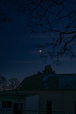 Hilal, ağaç dalları tarafından çerçevelenmiş ince bulutları olan koyu mavi bir gökyüzünde parlar. Aşağıda, eğimli çatısı olan mütevazı bir bina kırsal bir alanda oturuyor..