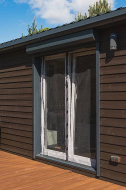 Koyu ahşap paneller, perdeli cam veranda kapıları, metal bir çatı ve parçalı bulutlu gökyüzünün altında ahşap bir güverte içeren bir kır evi..