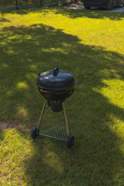 Tripod standı olan siyah bir kömür ızgarası çimenli bir çimenlikte oturur. Siyah bir araba bir çitin yanında kısmen görülebilir, açık hava ortamında yeşilliklerle çevrili..