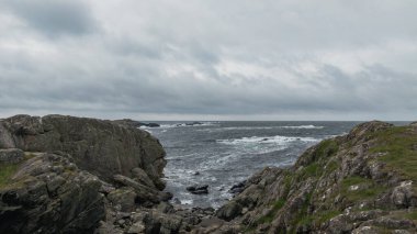 Kayalık kayalıklar ve pürüzlü arazi bulutlu bir gökyüzünün altında çalkantılı bir denizle buluşuyor. Yeşil çimenler karanlık sularla çelişiyor ve dramatik bir sahil manzarası yaratıyor..