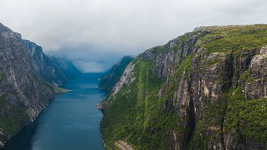 Norveç 'teki Lysefjord' un havadan görünüşü, yeşillikle kaplı engebeli uçurumlar, derin mavi bir fiyort, sis, alçak bulutlar ve üs boyunca dar bir yol..