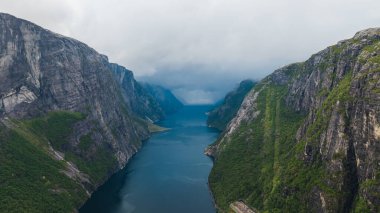 Norveç 'te Lysefjord' un hava perspektifi, sarp uçurumlar, yeşil, derin mavi sular ve alçak asılı bulutlar ile sisli bir sahne yaratıyor..