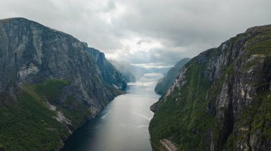 Norveç 'teki Lysefjord' un dramatik hava manzarası, dik uçurumlar, engebeli kaya yüzeyleri, sakin yansıtıcı sular ve atmosfer ekleyen bulutlu bir gökyüzü..