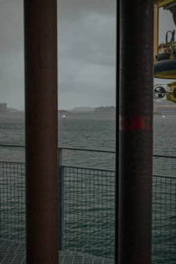 Norveç 'in Stavanger kenti yakınlarında bulutlu bir liman sahnesi, metal kutuplar, sarı endüstriyel bir yapı ve bulutlu bir gökyüzü altında uzak kara kütleleri yer alıyor..