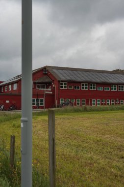 Çimlerle ve sarı çiçeklerle çevrili eğimli çatısı ve balkonu olan kırmızı ahşap bir bina. Bisikletler bulutlu bir gökyüzünün altında yakınlarda park halindedir..