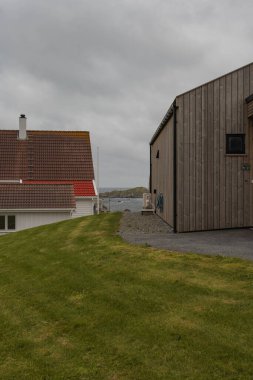 Biri kırmızı kiremit çatılı, biri ahşap cepheli iki bina denize bakan çimenli bir çimenlikte ve bulutlu bir gökyüzünün altındaki kayalık adalarda oturuyor..