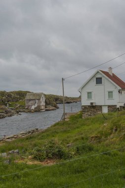 Norveç 'in Utsira Adası' ndaki sakin suların yakınındaki çimenli bir yamaçta, kanalın karşısında daha küçük bir bina bulunan beyaz ahşap bir ev bulunur..