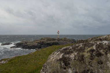 Deniz kenarındaki kayalık bir çıkıntıda kırmızı tepeli küçük bir deniz feneri dalgalı sularla, çimenli araziyle ve Utsira, Norveç 'te bulutlu bir gökyüzüyle çevrilidir..