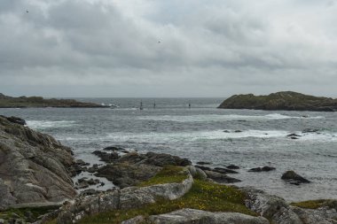 Çimenli kayalık arazi bulutlu bir gökyüzünün altında dalgalı bir denize çıkar. Norveç, Utsira yakınlarındaki sulardan küçük adalar ve seyrüsefer işaretleri yükseliyor..