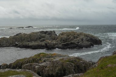 Kara yıpranmış kayalar, yosun parçaları ve bulutlu bir gökyüzü altında düşen dalgalarla kayalık kıyı şeridi. Muhtemelen Utsira ya da Stavanger, Norveç yakınlarında..