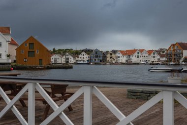 Renkli ahşap evler, rıhtımlı tekneler, beyaz parmaklıklı ahşap bir güverte ve bulutlu bir gökyüzünün yer aldığı Norveç 'in bir kasabasında sakin bir koy..