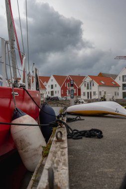 Görünür çamurlukları ve demirleme halatları olan kırmızı bir yelkenli rıhtımda duruyor. Yakınlarda beyaz ve kırmızı çatılı evler, üzerinde gri bulutlar olan Norveç fiyort kasabası.