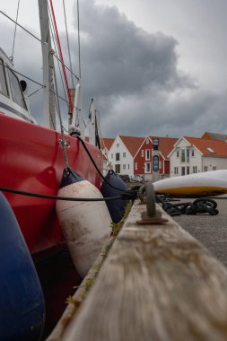 Görünür çamurlukları ve ipleri olan kırmızı bir yelkenli marinada dinleniyor. Turuncu kiremitli çatıları olan beyaz ve kırmızı ahşap evler bulutlu bir gökyüzünün altında arka planı çiziyor..