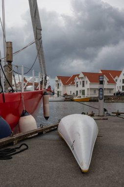 Kırmızı bir yelkenli, yakınlarda beyaz bir kanoyla bir rıhtıma demirlemiş. Kırmızı kiremitli çatıları olan beyaz evler Norveç 'te bulutlu bir gökyüzünün altında kıyı şeridi oluşturuyorlar..
